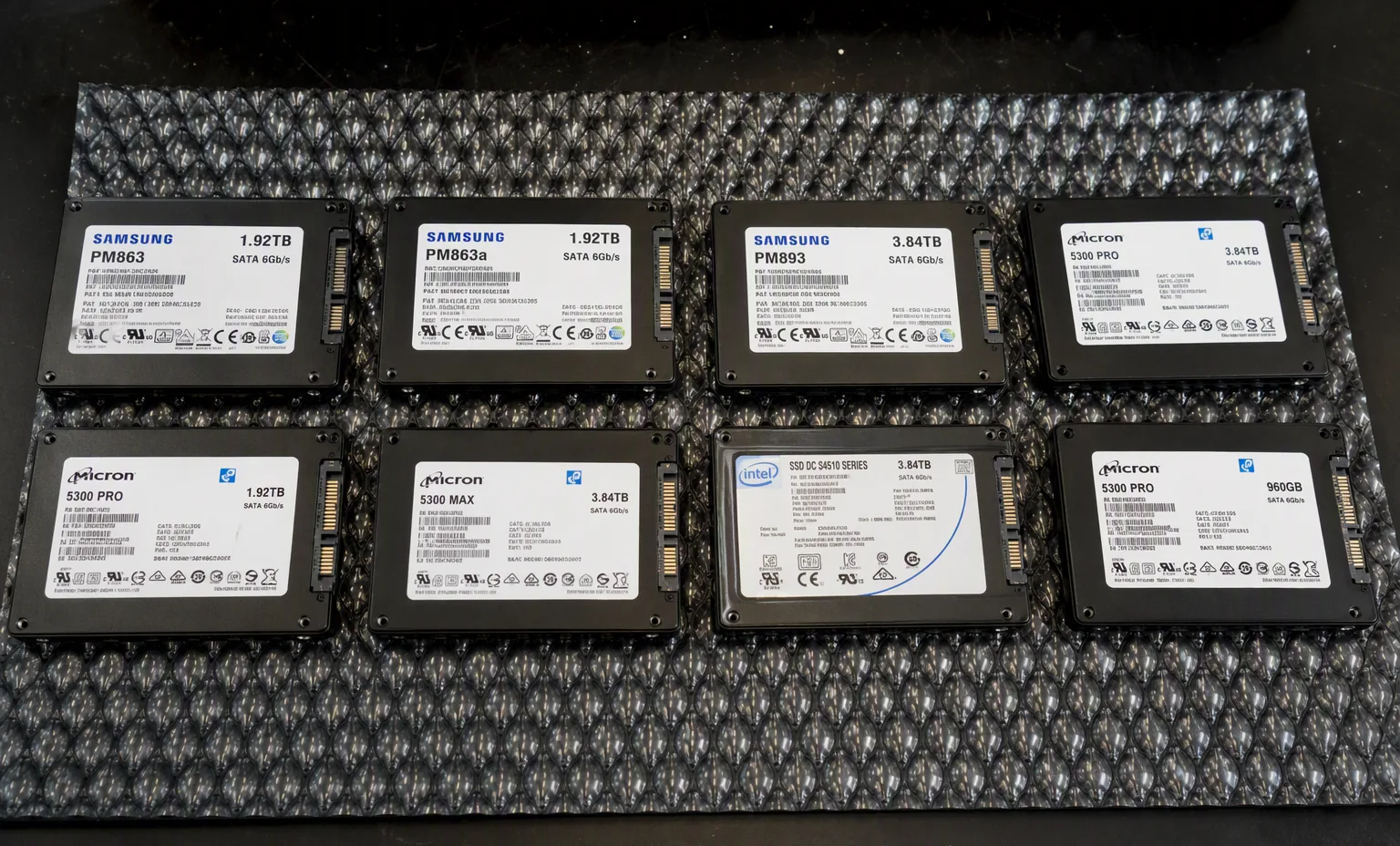 Photorealistic photograph of a row of eight mixed enterprise 2.5 inch SATA SSDs Samsung Intel Micron lined up on an anti-static mat for batch verification, dark lab bench.
